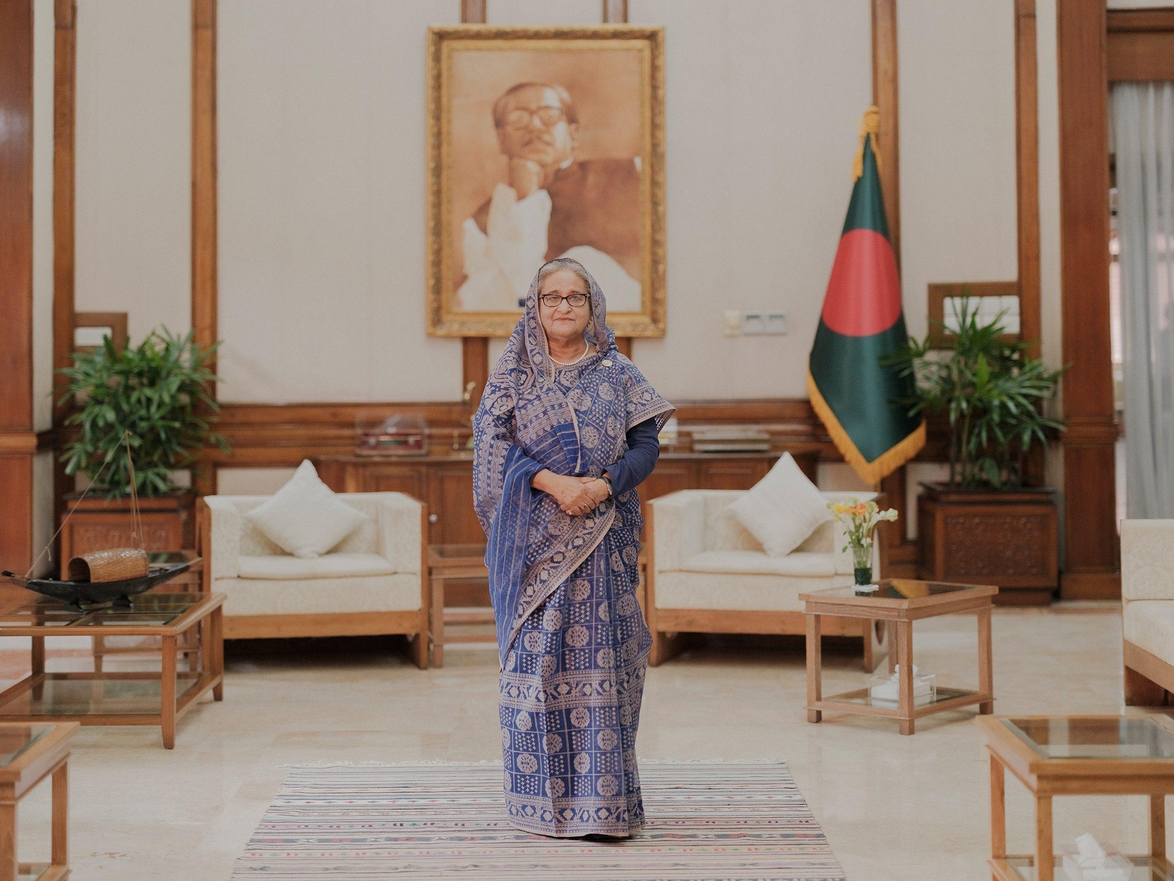 Sheikh Hasina standing in front of a portrait of Bangabandhu and the national flag